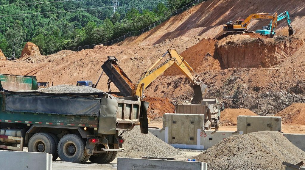 Cảnh thi công tại khu đầu đường dẫn vào suối Lương, dự án Vinhomes Làng Vân, với máy xúc đang bốc đất đá lên xe tải, phía sau là sườn đồi đã được đào xẻ, bao quanh bởi rừng cây xanh. 