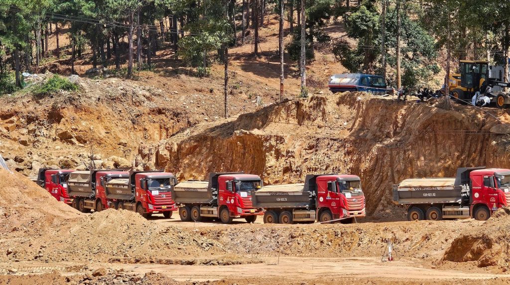 Dãy xe tải màu đỏ xếp hàng trên công trường dự án Vinhomes Làng Vân, chở đầy đất đá, phía sau là vách núi đã được đào xẻ và rừng cây bao quanh.