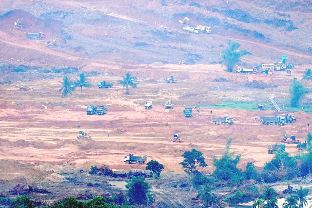 Toàn cảnh công trường dự án Vinhomes Làng Vân với nhiều xe tải, máy xúc và máy ủi đang san gạt đất, tạo hình mặt bằng, xen kẽ hàng dừa và cây xanh còn sót lại