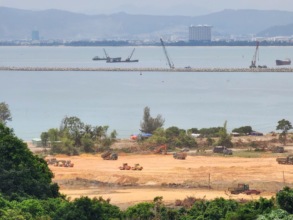 Cảnh thi công tại bãi biển dự án Vinhomes Làng Vân, với máy ủi, xe tải và máy xúc hoạt động, phía xa là cảng chắn sóng cùng tàu thuyền và đường chân trời của thành phố Đà Nẵng. 